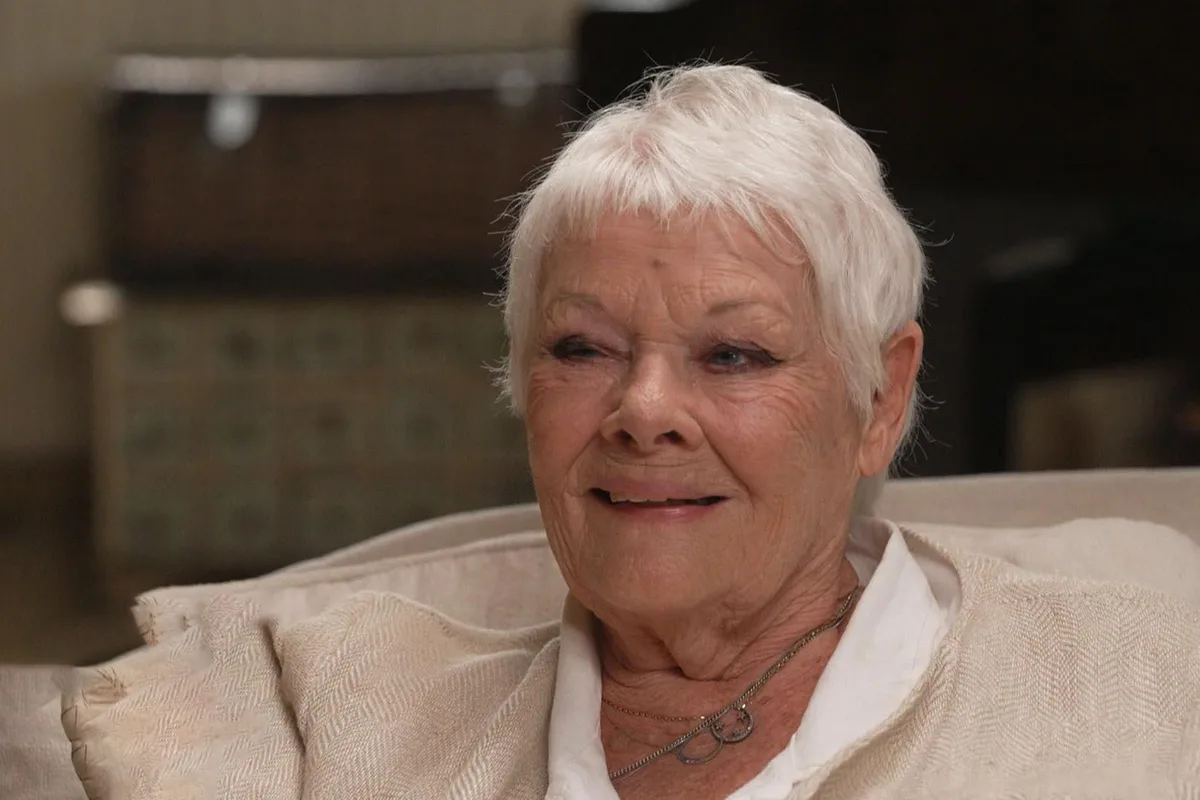 Judi Dench lors d'un entretien télévisé, assise dans un fauteuil clair et vêtue d'un haut blanc
